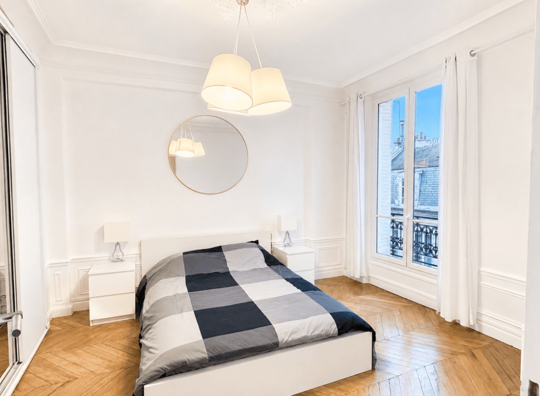 First bedroom with double bed, round mirror and French window onto a wrought-iron balcony — Le Marais · Paris 4e